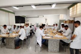 Estudantes estão em um laboratório, em dois grupos, sentados em duas mesas paralelas. Eles estão usando jaleco branco e parecem estar fazendo uma experiência química.