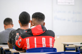 A imagem mostra três alunos de costas, todos homens de cabelos curtos e escuro. Eles estão sentados em uma fileira única, em uma sala de aula com um quadro branco e carteiras de encosto azul escuro.