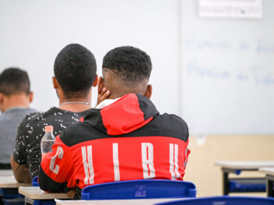 A imagem mostra três alunos de costas, todos homens de cabelos curtos e escuro. Eles estão sentados em uma fileira única, em uma sala de aula com um quadro branco e carteiras de encosto azul escuro.