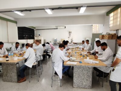 Estudantes estão em um laboratório, em dois grupos, sentados em duas mesas paralelas. Eles estão usando jaleco branco e parecem estar fazendo uma experiência química.