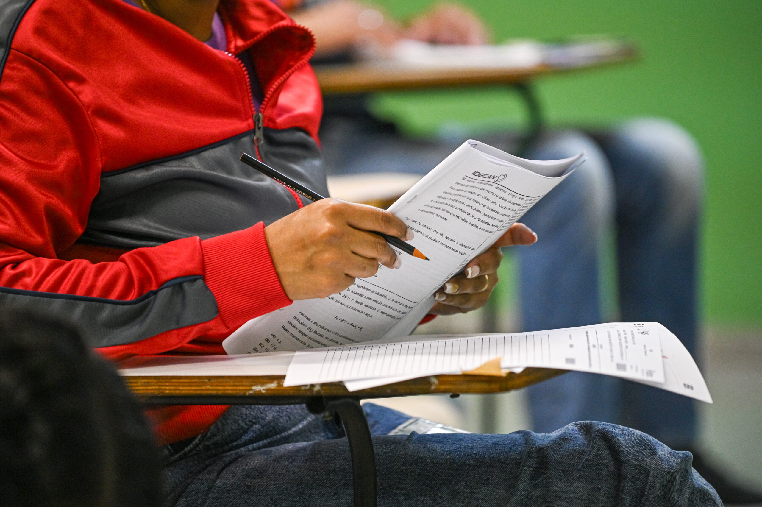 Close-up das mãos de uma pessoa sentada em uma carteira escolar, segurando um caderno de prova com o logotipo "CESGRANRIO". A mão direita, segurando uma caneta ou lápis, está posicionada sobre as questões. A pessoa veste uma jaqueta vermelha e cinza. Ao lado, na mesma carteira, há uma folha de respostas com códigos de barras. O fundo é desfocado, mostrando pernas de outras pessoas e uma parede verde.