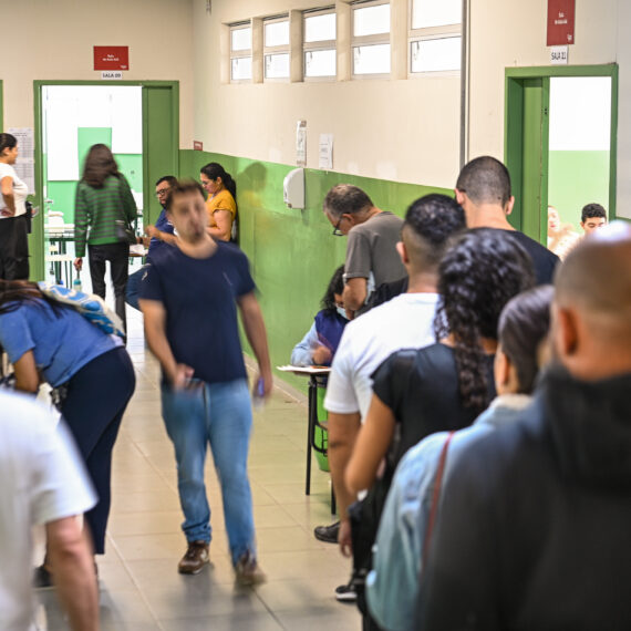 Um corredor movimentado com pessoas em fila de costas à direita. Outras pessoas caminham pelo corredor à esquerda, algumas desfocadas pelo movimento. O corredor tem paredes verdes e brancas, portas verdes e janelas altas na parede direita. Placas vermelhas indicam números de salas.