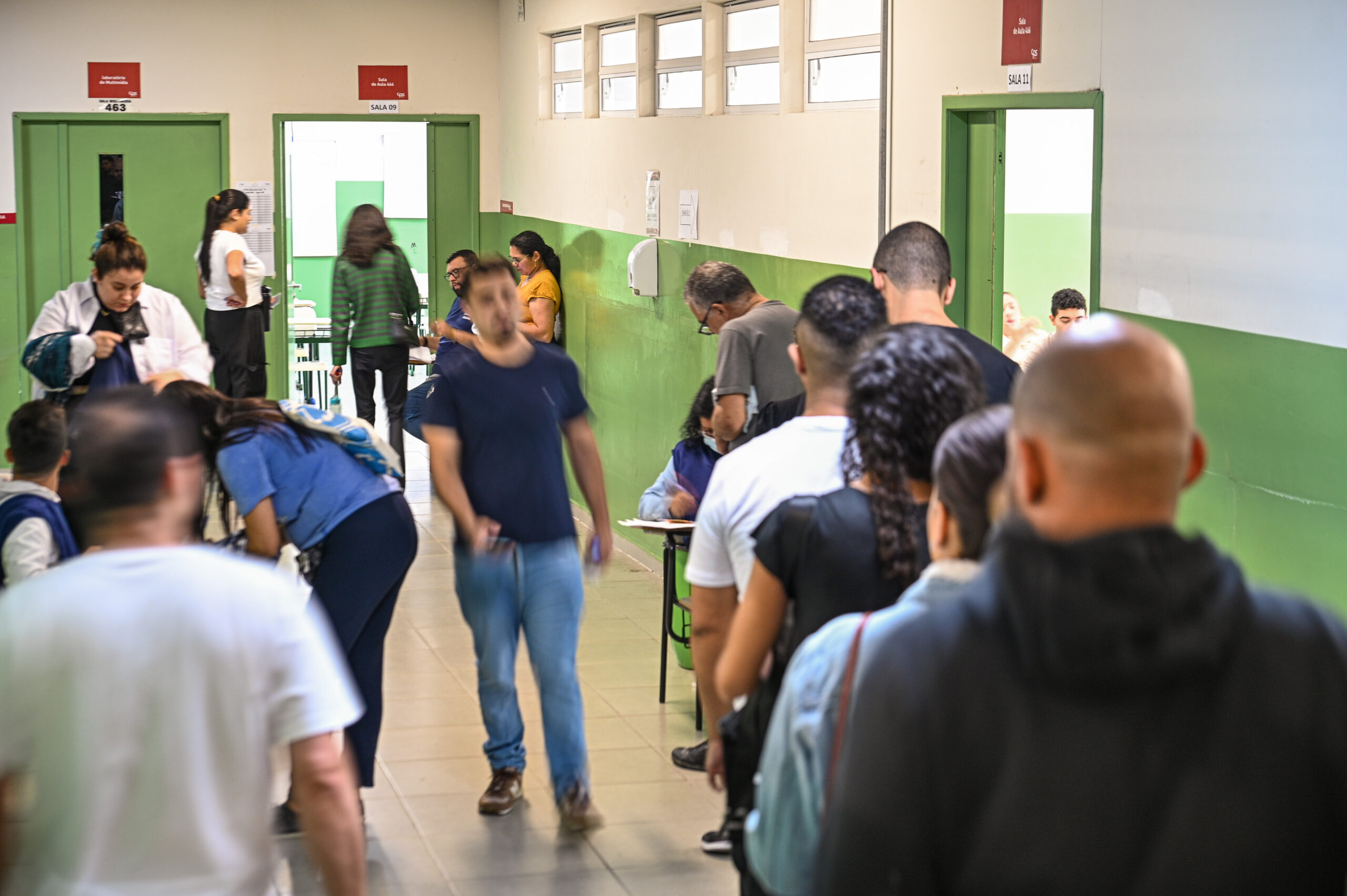 Um corredor movimentado com pessoas em fila de costas à direita. Outras pessoas caminham pelo corredor à esquerda, algumas desfocadas pelo movimento. O corredor tem paredes verdes e brancas, portas verdes e janelas altas na parede direita. Placas vermelhas indicam números de salas.