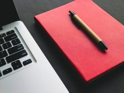 computador com caderno vermelho e caneta ao seu lado